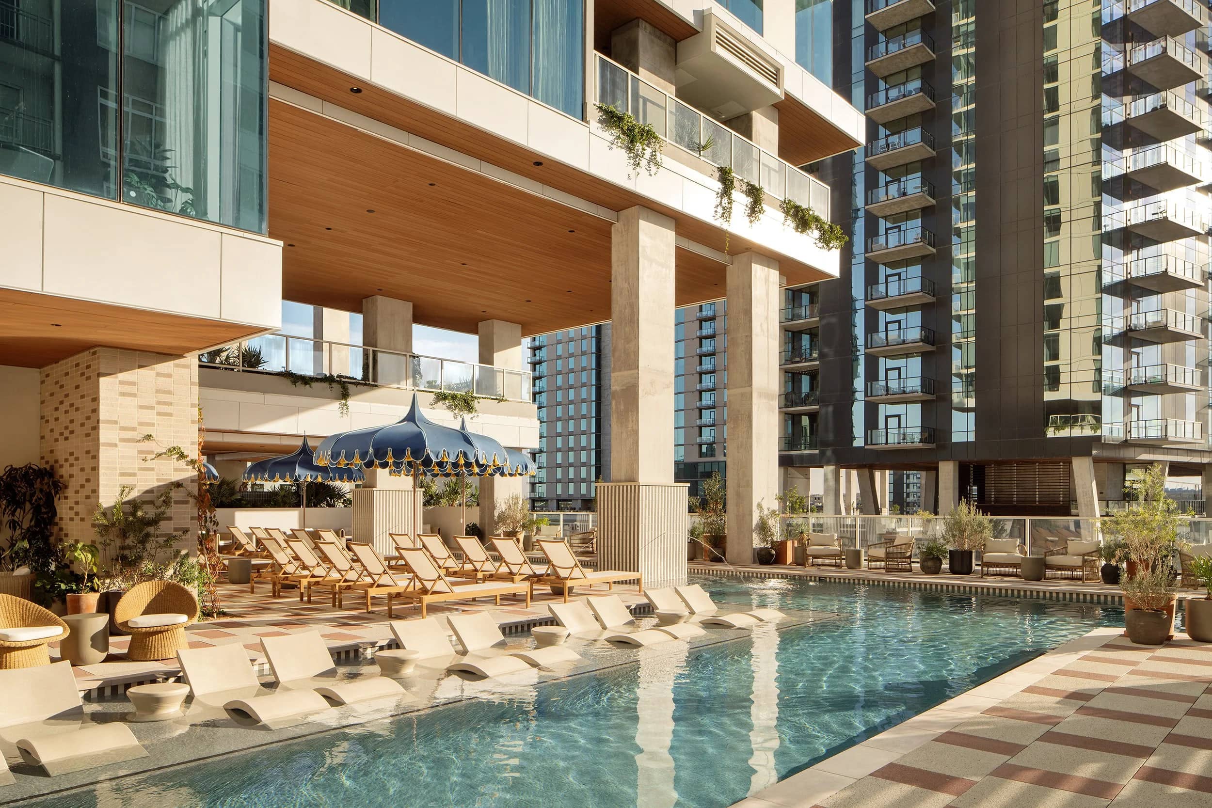 A pool terrace at golden hour, loungers and scalloped umbrellas along the water, with residential towers behind.