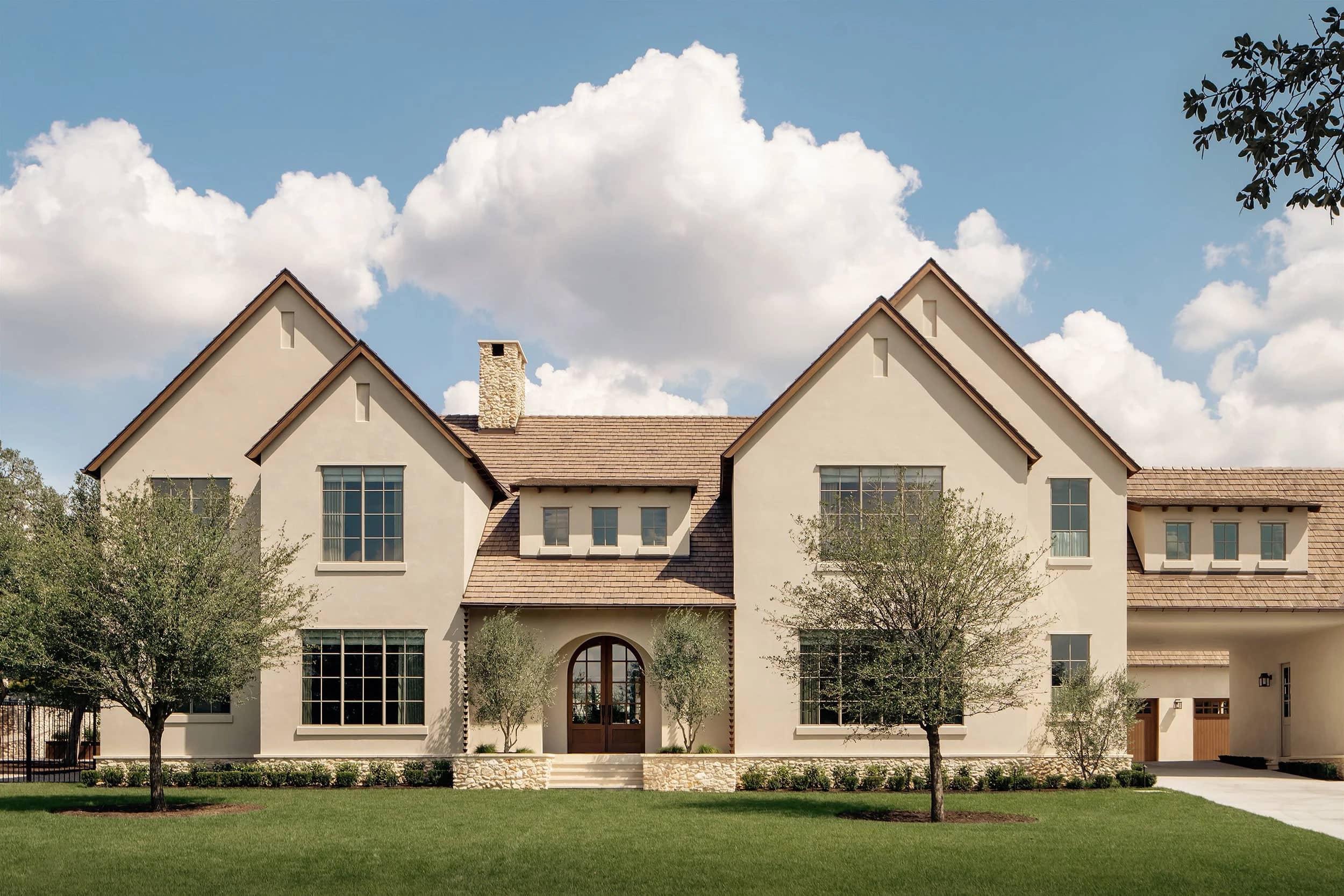 A limestone and stucco residence with steep gables, a cedar shingle roof, and olive trees along the front elevation.